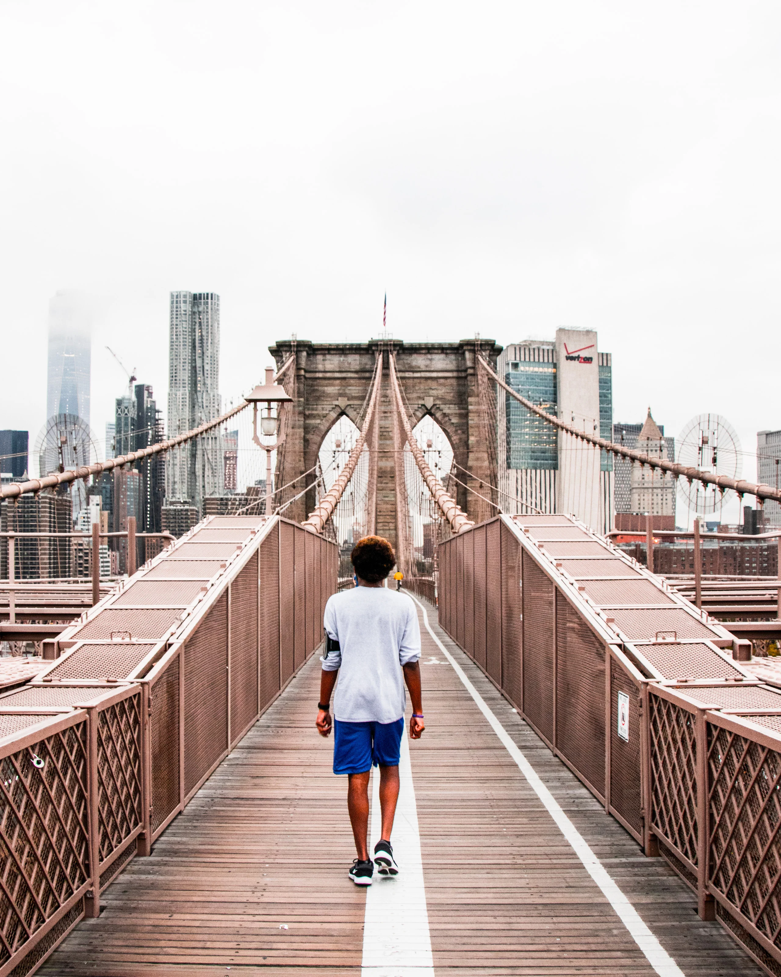 A man walking the Brooklyn Bridge promenade towards the stone tower and a misty skyline